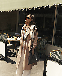 A woman in a trench coat stands in front of a cafe with tables and chairs visible in the background. carousel Product Image 5