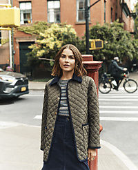 A woman in a quilted jacket stands on a sidewalk with a blurred city street and a cyclist in the background. carousel Product Image 3