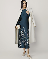 A woman in a teal dress with white floral embroidery and a white coat stands against a plain white background, holding a white clutch. carousel Product Image 3