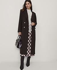 A woman in a long brown coat and plaid skirt stands against a plain white background, holding a dark handbag. carousel Product Image 2