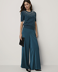 A woman in a teal embellished sweater and wide-leg pants poses against a plain, light background, holding a black clutch. carousel Product Image 1
