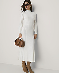 A woman in a white cable-knit dress and brown boots stands against a plain white wall, holding a brown handbag. carousel Product Image 1