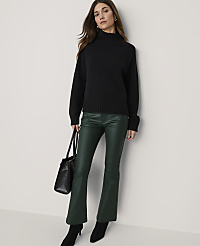 A woman in a black turtleneck and green flared pants stands against a plain, light-colored wall, holding a black handbag. carousel Product Image 1
