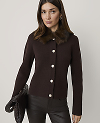 A woman in a brown cardigan with a fur collar and dark pants holds a clutch in the foreground against a plain background. carousel Product Image 1