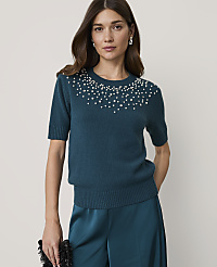 A woman in a teal sweater adorned with pearls stands against a plain, light background, holding a black clutch. carousel Product Image 1
