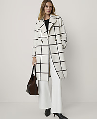 A woman in a white and black plaid coat and white pants stands in front of a plain white background, holding a brown purse. carousel Product Image 1