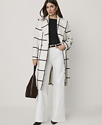 A woman in a cream and black checkered coat and white flared jeans stands against a plain white background, holding a brown bag. carousel Product Image 1