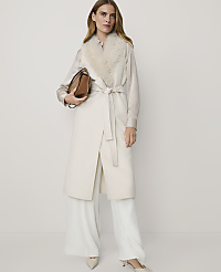A woman in a cream coat with a fur collar and wide-leg pants stands against a plain, light background, holding a brown handbag. carousel Product Image 1