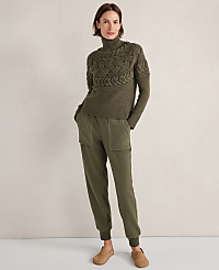 A woman in a green cable-knit turtleneck sweater and matching ribbed pants stands against a plain, light brown background. carousel Product Image 1