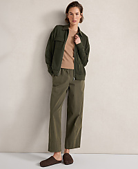A woman in a tan shirt, green jacket, and olive pants stands against a textured cream wall, wearing brown slippers. carousel Product Image 3