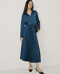 A woman in a teal satin blouse and pleated skirt stands against a plain white background, holding a black clutch. carousel Product Image 1