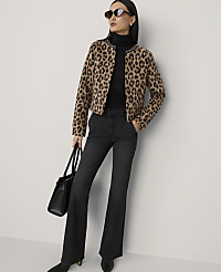 A woman in a leopard print jacket and dark flared jeans stands against a plain, light-colored wall, holding a black handbag. carousel Product Image 4