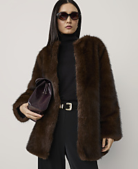 A woman in a luxurious brown fur coat and black turtleneck stands against a plain, light gray background, holding a dark purple clutch. carousel Product Image 1