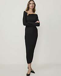 A woman in a black off-the-shoulder ribbed knit top and matching skirt stands against a plain, light-colored background.