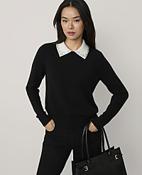 A woman wearing a black sweater with a beaded white collar and black pants holds a black handbag against a plain light gray background. carousel Product Image 1