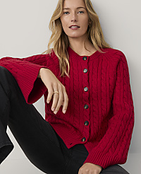 A blonde woman wearing a red cable-knit cardigan and black pants sits against a plain white background. carousel Product Image 2
