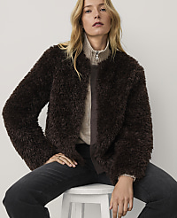 A woman wearing a dark brown faux-fur jacket and black jeans sits on a white stool against a plain light-colored background. carousel Product Image 4