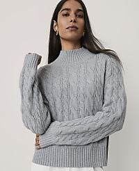 A woman wearing a grey cable-knit sweater stands against a plain, light-colored background. carousel Product Image 1