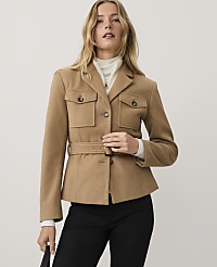 A woman wearing a tan belted jacket, white turtleneck, and black trousers stands against a plain light gray background. carousel Product Image 1