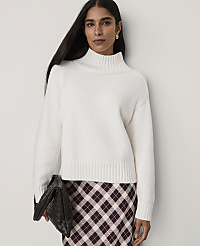 A woman wearing a white mock-neck sweater and a plaid skirt stands against a plain, light-colored wall. carousel Product Image 1