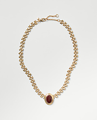 A gold chain necklace with a red oval gemstone pendant rests on a plain light gray background. carousel Product Image 1