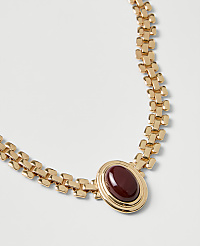 A gold chain necklace with a red oval gemstone pendant rests on a plain white background. carousel Product Image 2