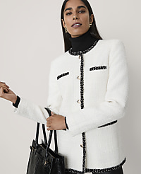 A woman wearing a white tweed jacket and black turtleneck holds a black handbag against a plain, light-colored background. carousel Product Image 1