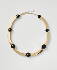 A gold-toned necklace with black spherical beads rests on a plain light gray background. carousel Product Image 1