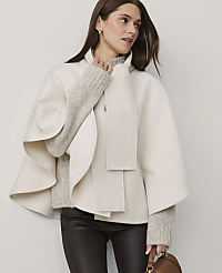 A woman wearing a cream-colored cape and brown leather pants stands against a plain, light-grey background. carousel Product Image 1