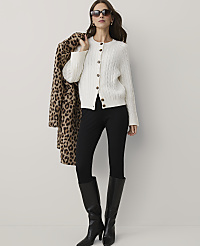 A woman wearing a white cable-knit cardigan, black pants, and black boots stands against a plain white background while holding a leopard-print coat over her shoulder. carousel Product Image 1