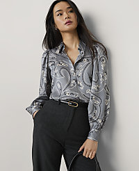 A woman wearing a grey paisley print blouse and dark trousers stands against a plain light grey wall. carousel Product Image 1