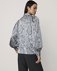 A woman wearing a grey paisley print blouse and dark trousers stands against a plain light grey background. carousel Product Image 2