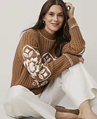 A woman wearing a brown patterned cable-knit sweater and white trousers sits against a plain light gray background. carousel Product Image 3