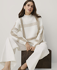 A woman wearing a white and beige patterned sweater and white trousers sits on a dark wooden block against a plain light grey background. carousel Product Image 3