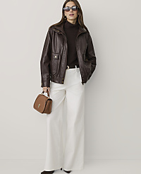 A woman wearing a brown leather jacket, dark turtleneck, and wide-leg white trousers stands against a plain, light-colored background. carousel Product Image 1