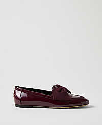 A burgundy patent leather flat shoe with a bow sits in the foreground against a plain, light gray background. carousel Product Image 1