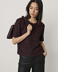 A woman wearing a red and black tweed top and black leather pants poses with a burgundy handbag against a plain light grey background. carousel Product Image 1
