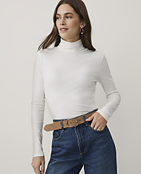 A woman wearing a white turtleneck and blue jeans with a brown belt stands against a plain, light-colored background. carousel Product Image 1