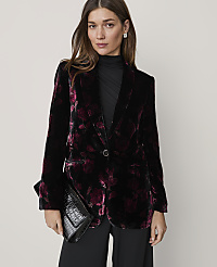 A woman wearing a floral velvet blazer and black clothing holds a black clutch against a plain, light-colored background. carousel Product Image 1