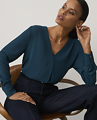 A woman wearing a teal V-neck blouse and dark blue jeans sits in a wooden chair against a plain, light-colored background. carousel Product Image 1