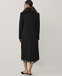 A woman stands with her back to the camera wearing a long black coat with a brown faux-fur collar against a plain, light-colored background. carousel Product Image 2