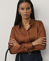 A woman with long dark hair wearing a copper-colored satin shirt and dark blue jeans stands with her arms crossed against a plain light gray background. carousel Product Image 1