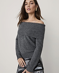A woman wearing a grey off-the-shoulder sweater and holding a black clutch stands against a plain, light-colored background. carousel Product Image 1