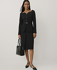 A woman wearing a black blazer, skirt, and heels stands holding a black handbag against a plain white background. carousel Product Image 1