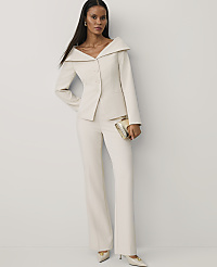 A woman wearing a cream-colored tailored suit and holding a gold clutch stands against a plain white wall. carousel Product Image 3
