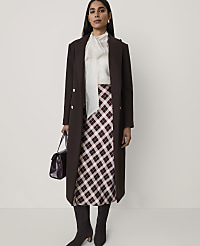 A woman wearing a long brown coat, a white blouse, and a plaid skirt stands against a plain, light-colored background. carousel Product Image 1