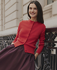 A smiling woman wearing a red cardigan and a dark purple skirt leans against an ornate black iron railing in front of a light-colored stone building. carousel Product Image 3