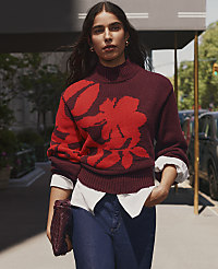 A woman wearing a maroon floral sweater and blue jeans stands on a city sidewalk with cars and greenery in the background. carousel Product Image 4