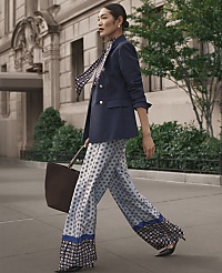 A woman in a navy blazer and patterned wide-leg trousers walks along a city sidewalk in front of a classical stone building and green foliage. carousel Product Image 4