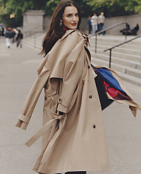 A woman in a tan trench coat stands in the foreground against a blurred city background with stone steps and pedestrians. carousel Product Image 4